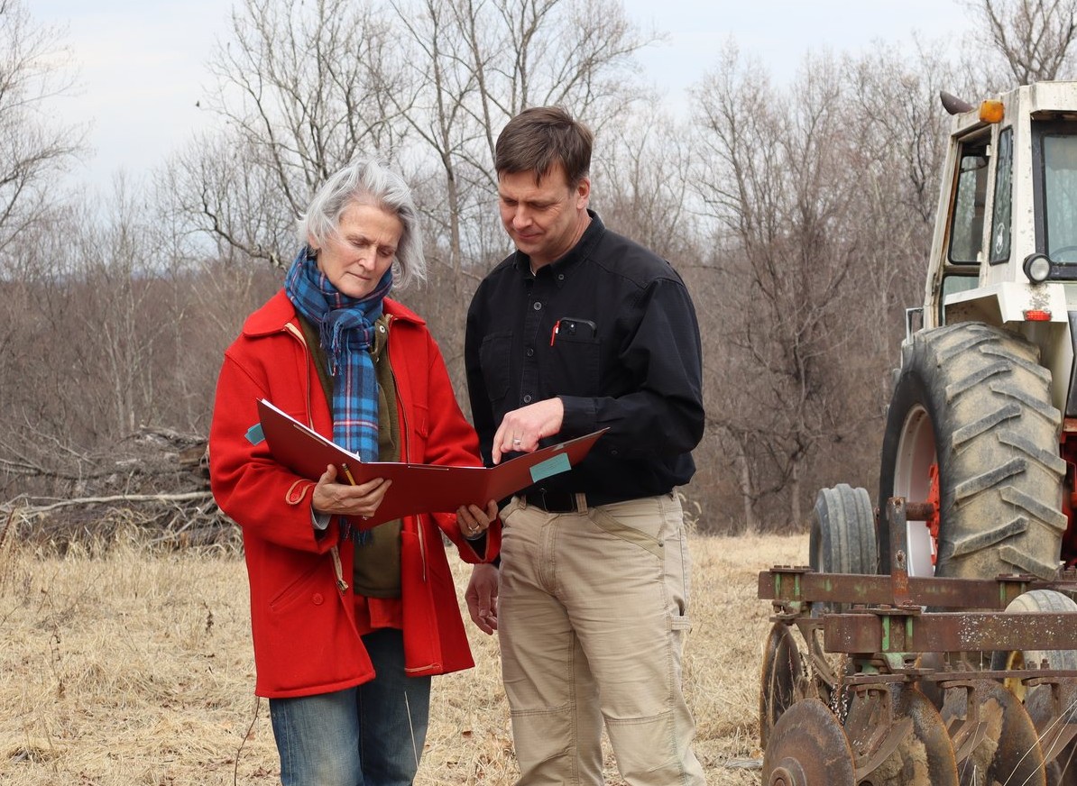 John Kluthe, of KES LLC, reviewing a conservation plan in the field with a client farmer beside farm equipment (heavy disk) and tractor.